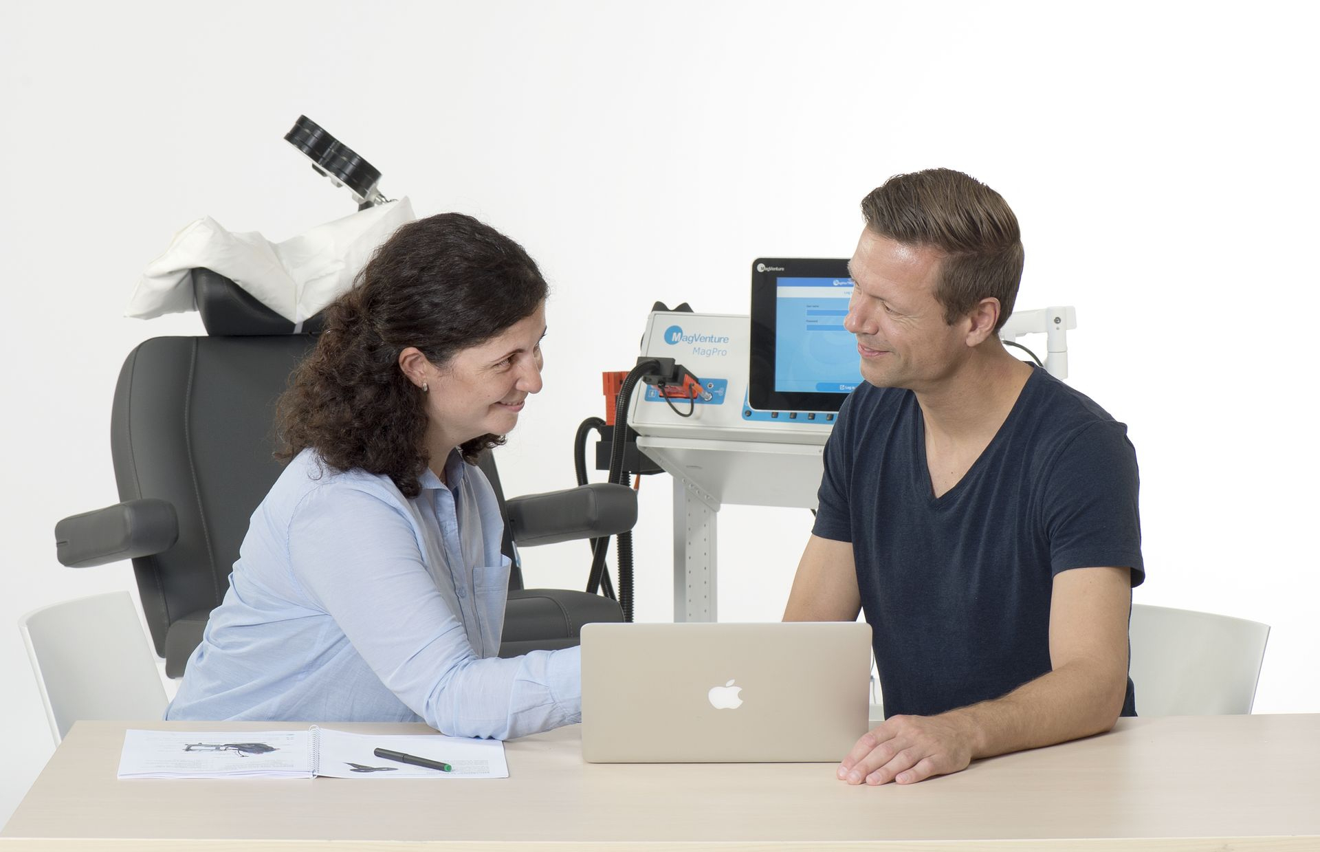 Doctora y paciente sentados en la mesa frente a un ordenador portátil y con una máquina de estimulación magnética transcraneal de fondo